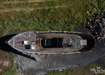 An aerial view of an old, weathered boat resting on a rocky shoreline, surrounded by green grass and scattered pebbles. The boat's deck shows signs of rust and decay, with an empty space where equipment might have once been. Various tools and objects are scattered around the boat, adding to the scene of abandonment.