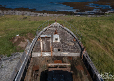 An abandoned boat sits on a grassy area near the coastline, with a partially visible rusted railing and worn wooden deck. The boat's bow is angled toward the water, showing signs of decay, including peeling paint and a damaged door. In the background, the blue sea meets a rocky shoreline with patches of seaweed visible in shallow water. The scene conveys a sense of neglect and tranquility, surrounded by nature.