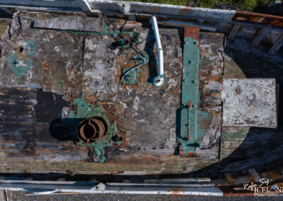 An aerial view of a weathered wooden boat deck, showcasing a mix of peeling paint in shades of green and rusted metal parts. The deck is marked by textures of aged wood and metal, with a circular rusted pipe and various mechanical components partially visible. The surrounding area appears to have some grassy elements, suggesting an outdoor setting.