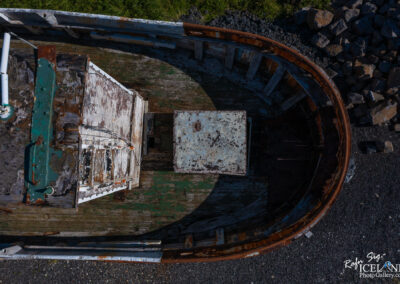 An aerial view of an old, weathered fishing boat resting on gravel. The boat features peeling paint, rusted metal, and a wooden deck showing signs of decay. In the center, there is a metal lid or box, and the surrounding area is dotted with small, irregularly shaped stones and patches of grass.