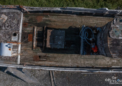 An aerial view of an old, weathered fishing boat is shown, featuring a wooden deck with patches of green moss and rust. The deck is cluttered with various objects, including a metal hatch, circular vents, and tubes, while remnants of peeling paint and corrosion add to the boat's neglected appearance. Tall grass and a gravel surface are visible around the boat, suggesting it is no longer in active use.