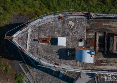 An aerial view of an old, weathered boat resting on a rocky shore. The boat's bow is visible, showcasing its worn wooden surface, patches of peeling paint, and rusted metal fixtures. Surrounding the boat, there is green grass and some rocky terrain. The sunlight casts shadows on the deck, highlighting its aged condition.