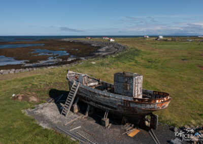 An abandoned wooden boat rests on a grassy shore, with a staircase leading up to its deck. It has rusted sections and weathered paint, indicating years of exposure to the elements. In the background, the coastline features scattered rocky formations and patches of vegetation, with several small, colorful houses visible on the horizon. The sky is clear and blue, framing the tranquil seascape.