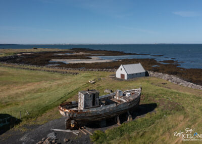 A rusted, abandoned fishing boat sits on a grassy shore near the water, with a simple white shed in the background. The landscape features rocky beach areas and patches of seaweed along the shoreline, under a clear blue sky. The scene conveys a sense of tranquility and the passage of time in a coastal environment.