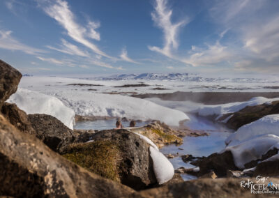 A serene natural hot spring surrounded by snowy landscapes and rocky formations, under a clear blue sky with wispy clouds. Two people can be seen relaxing in the warm water, creating a peaceful atmosphere in the stunning winter scenery. The distant mountains add to the breathtaking backdrop of this tranquil setting.