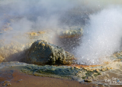 A geothermal area with steam rising from the ground, highlighting a bubbling hot spring. Rocks and mineral deposits are visible, showcasing shades of blue, green, and orange. Water is splashing out from a central point, creating a dynamic and lively scene.