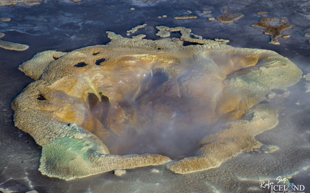 A close-up view of a colorful geothermal feature, resembling a bubbling hot spring surrounded by textured mineral deposits. The center shows steam rising from the water, while the edges display shades of yellow, green, and brown, indicating the presence of various minerals. The ground surrounding the spring is dark and appears moist, adding to the dynamic and vibrant appearance of this natural phenomenon.