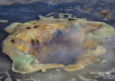 A close-up view of a colorful geothermal feature, resembling a bubbling hot spring surrounded by textured mineral deposits. The center shows steam rising from the water, while the edges display shades of yellow, green, and brown, indicating the presence of various minerals. The ground surrounding the spring is dark and appears moist, adding to the dynamic and vibrant appearance of this natural phenomenon.
