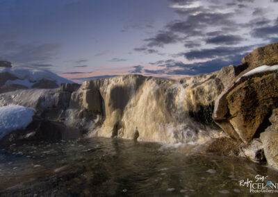 A serene scene featuring a flowing waterfall cascading over a rocky ledge into a clear pool of water, surrounded by smooth stones. The water is shimmering, with splashes creating soft ripples on the surface. In the background, a sky filled with clouds transitions from light orange to deep blue, indicating either sunrise or sunset, adding a warm glow to the landscape. Snow drapes over some of the rocks, contrasting with the darker, wet surfaces below.