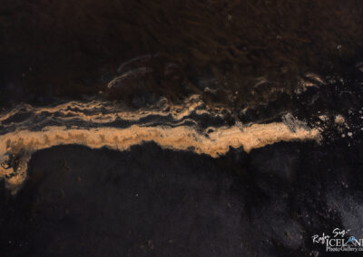 An aerial view of a beach scene, featuring a stark contrast between dark black sand and a wavy line of golden-brown sand, creating a natural decorative pattern. The textures and colors highlight the beauty of the coastline, with gentle waves subtly reflecting light.