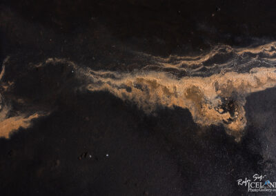 An artistic close-up of a textured surface featuring contrasting shades of dark black and sandy gold. The image captures a swirling pattern where the dark ground meets lighter particles, creating a natural abstract design. The composition highlights the interplay of colors and textures, evoking a sense of movement and fluidity.
