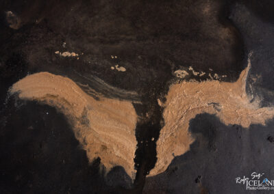An aerial view of a sandy area with dark wet surfaces. The sand is arranged in a flowing pattern, contrasting against the dark background, creating an abstract design that resembles wings or flowing shapes. Small patches of foam and lighter sands are scattered across the surface, adding texture and complexity to the scene.