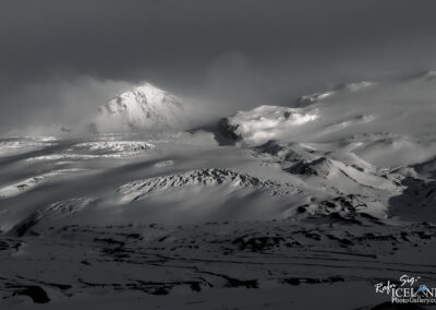 A dramatic black-and-white landscape featuring a towering snow-covered mountain partially obscured by clouds. The foreground showcases rolling hills and undulating snowdrifts, creating a sense of depth and texture. The sky is gray and moody, adding to the atmospheric effect of the scene.