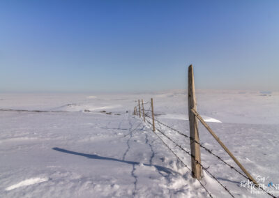 A vast, snow-covered landscape stretches out under a clear blue sky. In the foreground, a wooden fence runs parallel to the viewer, with barbed wire strung between the posts. The snow is undisturbed, creating a smooth surface, with faint shadows from the fence stretching across it. In the distance, gentle hills can be seen rising from the flat expanse of snow. The overall scene conveys a sense of tranquility and isolation in a wintry environment.