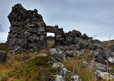 A partially collapsed stone structure stands amid rugged terrain, with scattered rocks and patches of grass surrounding it. The sky above is overcast, enhancing the somber and historical mood of the landscape. The stones are unevenly stacked, indicating age, while tufts of grass and moss add a touch of natural vibrancy to the scene.