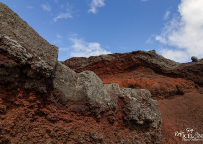 A landscape featuring rocky formations with varying textures and colors, including gray and reddish hues. The sky above is bright blue with scattered clouds, creating a contrast with the earthy tones of the rocks. The image captures a section of the terrain, highlighting the natural geological structure.