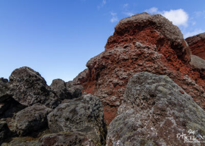 A close-up view of textured volcanic rocks in various shades of grey and red, set against a clear blue sky with a few clouds. The rocks exhibit moss and lichen growth, showcasing the natural landscape's rugged and colorful geology.