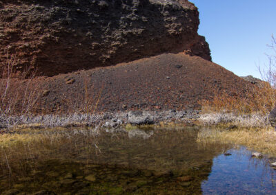 A rocky, volcanic cliff rises sharply against a clear blue sky, with patches of green moss on top. In the foreground, a shallow pool of water reflects the cliff and surrounding dry grass. The ground is scattered with small rocks and pebbles, indicating a rugged terrain.