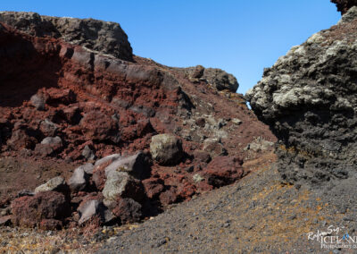 A rocky volcanic landscape featuring red and dark grey lava formations, rugged terrain, and a clear blue sky. The foreground consists of various sizes of lava rocks and a sloping surface, with a rocky outcrop in the background highlighting the natural textures and colors of the volcanic material.