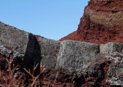A close-up view of rocky terrain, featuring large, rugged gray rocks covered with patches of lichen or moss, set against a backdrop of reddish volcanic soil and formations. The clear blue sky is visible in the background, enhancing the contrast between the earth tones. Sparse vegetation in the foreground adds an organic touch to the scenery.