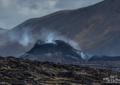 A volcanic landscape featuring a small erupting volcano, emitting wisps of smoke against a backdrop of rugged, mountainous terrain. The foreground showcases dark, rocky lava formations scattered with patches of moss, while the sky above is partially cloudy, creating an atmospheric scene.
