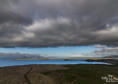 A panoramic view of a serene landscape featuring a calm lake surrounded by green hills and rocky terrain. The sky is partly cloudy, with dark clouds looming above while patches of blue peek through. In the distance, islands and hills can be seen, contributing to the tranquil natural scenery. A winding dirt path is visible leading into the foreground.