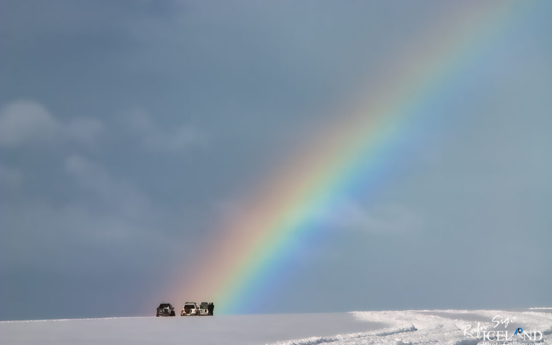 Two vehicles parked on a snowy landscape, with a vibrant rainbow arcing across the sky in the background. The scene captures a calm, serene atmosphere under a cloudy sky.