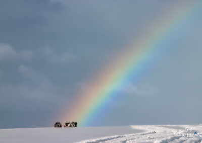 Two vehicles parked on a snowy landscape, with a vibrant rainbow arcing across the sky in the background. The scene captures a calm, serene atmosphere under a cloudy sky.