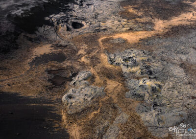 An aerial view of an expansive volcanic landscape featuring a mix of dark lava rock and patches of lighter earthy tones. The terrain is characterized by rugged and uneven surfaces, with winding pathways and sparse vegetation intermingled among the rock formations. Some areas appear smoother while others display a cracked and rough texture, suggesting a dramatic geological history. The scene is illuminated by natural light, highlighting the variations in color and texture across the landscape.