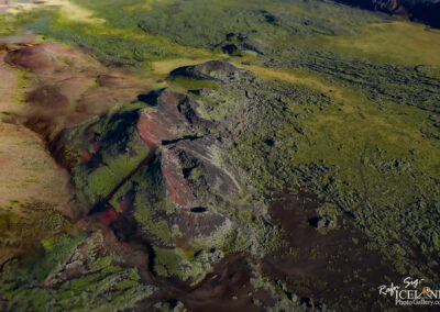 An aerial view of a rugged landscape featuring rolling hills covered in a mix of green vegetation and dark volcanic rock. The terrain shows various textures and colors, with patches of reddish-brown earth visible among the greenery. There are also traces of lava flow and rocky formations that hint at a volcanic origin. The sky above is partially obscured by the expansive terrain, showcasing the natural beauty of the area.