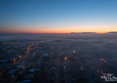 Aerial view of a town partially enveloped in fog during twilight, with soft hues of orange and blue in the sky. Streetlights and building outlines are visible through the mist, creating a serene and atmospheric scene. Mountains are faintly visible in the background.