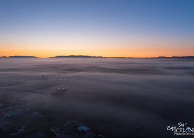 A breathtaking aerial view of a foggy landscape at dawn, showcasing a smooth blanket of mist covering the ground below. In the distance, silhouettes of mountains can be seen against a gradient sky transitioning from deep blue to warm orange hues. Subtle lights from structures peeking through the fog create a serene atmosphere, highlighting the quiet beauty of the early morning.
