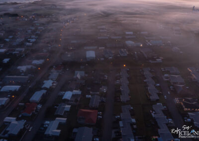 An aerial view of a foggy residential area at dusk. The scene captures rows of houses with a mix of colors in their rooftops, including grey, red, and white. Street lights are visible, casting a warm glow amid the mist, while some vehicles are parked along the roads. The background features blurred outlines of structures and trees partially obscured by the fog.