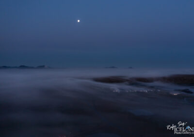 A serene, twilight landscape featuring a hazy expanse with low-lying fog enveloping the terrain. In the sky above, a bright full moon casts a gentle light, illuminating the scene. Silhouetted hills and mountains emerge faintly in the background, blending into the soft blue and gray tones of the evening sky. The atmosphere conveys a sense of tranquility and calm.
