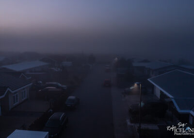 A foggy street scene at dusk, with dim lighting illuminating a quiet residential area. Several houses with sloped roofs are visible, some adorned with carports. The road is lined with parked cars, and a few glowing streetlights cast a soft light amid the haze, creating a calm yet eerie atmosphere. The background fades into a thick fog, obscuring any further details.