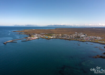 Aerial view of a coastal town located on the edge of a small peninsula with a harbor. The town features a mix of residential and commercial buildings, with red and white-roofed structures, surrounded by a serene blue water landscape and green fields. In the background, you can see distant mountains under a clear sky, enhancing the picturesque scenery. The shoreline includes a breakwater and gentle waves lapping at the rocks.