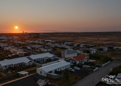 Aerial view of a residential area with various houses and rooftops, set against a backdrop of a sunset. The sun is partially visible, casting orange and pink hues in the sky. In the distance, there are fields and a faint outline of mountains. The scene conveys a serene, picturesque atmosphere of a quiet neighborhood at dusk.