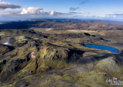 A panoramic aerial view of a rugged, mountainous landscape featuring various shades of green and brown. The terrain includes rocky outcrops, rolling hills, and a serene blue lake nestled among the hills. The sky above is partly cloudy, with soft white clouds drifting amidst a bright blue backdrop. In the distance, additional mountain ranges can be seen, fading into the horizon.