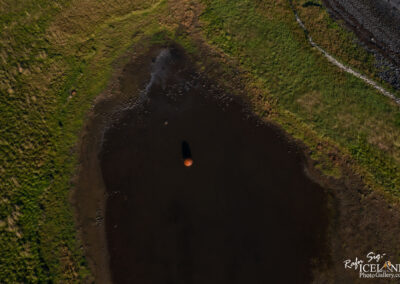 An aerial view of a dark, circular body of water surrounded by lush green grass and patches of brown land. A solitary orange object floats on the water's surface, casting a shadow. The edges of the water feature taper off into rocky and grassy areas, creating a natural contrast in textures and colors.