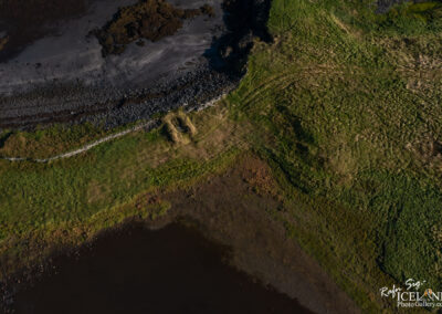 An aerial view of a coastal landscape featuring a mix of dark sandy beach and rocky shoreline. Lush green grass and various earth tones emerge where the land meets a body of water, which appears dark and reflective. A low stone wall is visible, and patches of brown grass intersperse the green areas, highlighting the natural textures of the terrain.
