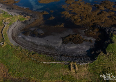 Aerial view of a rocky shoreline merging with patches of wetland and sea. The scene features a curving shoreline with a mix of dark sand, pebbles, and patches of green grass. Calm water is visible, with some seaweed and small islands of rock. A stone structure is partially visible in the greenery near the shore, enhancing the natural landscape.