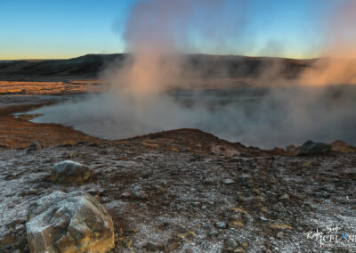 A geothermal landscape featuring steam rising from a hot spring amidst rocky terrain. The ground displays a mix of earth tones with patches of frost, and distant hills are visible under a clear blue sky at dawn. The scene conveys a sense of natural beauty and geothermal activity.