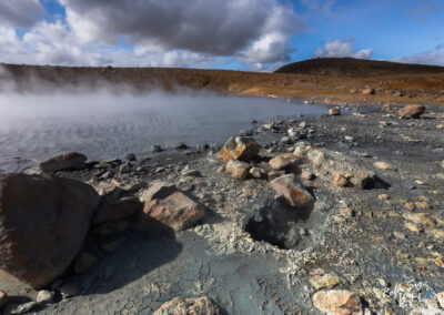 A geothermal landscape featuring a steamy, gray, mineral-rich hot spring surrounded by rocky terrain. The ground is uneven with various sizes of stones scattered around, and a mist rises from the water's surface under a partly cloudy sky. In the distance, a barren hillside outlines the scene, creating a contrast with the vibrant colors of the rocks and steam.