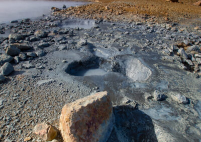 A geothermal landscape featuring a rocky terrain with patches of gray mud and steaming water. In the foreground, there are large stones and irregular depressions filled with water. The background shows a barren, hilly area under a partly cloudy sky, creating a stark contrast between the earthy tones of the landscape and the blue hues of the water and sky.