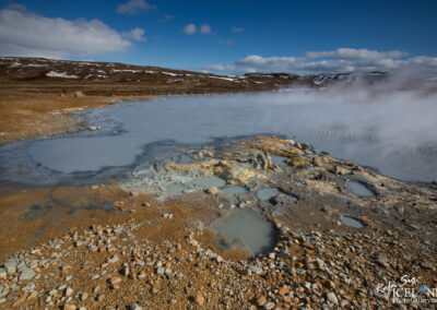 A vast geothermal landscape featuring a steaming, bluish-gray pool surrounded by rocky terrain. The ground is a mix of earth tones with patches of brown, gray, and yellow minerals. Wisps of steam rise from the water, contrasting with a clear blue sky dotted with clouds. In the background, a rugged, snow-capped hillside adds depth to the scene.