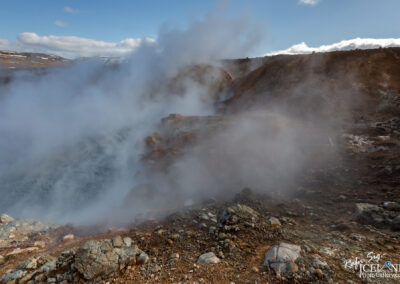 A geothermal landscape featuring a rocky terrain with steam rising from the ground. The scene is partially obscured by white steam, creating a misty atmosphere. The background includes a distant mountainous area under a blue sky with scattered clouds. The ground is composed of various shades of brown and grey rocks, suggesting volcanic activity.