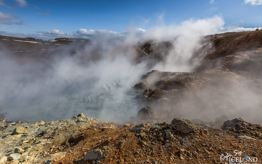 A rugged geothermal landscape featuring steam rising from a shallow hot spring, surrounded by rocky terrain. The scene is set under a blue sky with scattered clouds, and hints of snow are present on the distant hills. The ground is a mix of earth tones, showcasing rocky outcrops and a combination of textures from the geothermal activity.