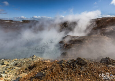 A rugged geothermal landscape featuring steam rising from a shallow hot spring, surrounded by rocky terrain. The scene is set under a blue sky with scattered clouds, and hints of snow are present on the distant hills. The ground is a mix of earth tones, showcasing rocky outcrops and a combination of textures from the geothermal activity.