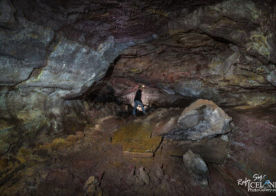 A dimly lit Lava cave filled with large, uneven rock formations and a mix of dirt and gravel on the ground. A person is seen walking on the rocky terrain, wearing a headlamp, and the cave walls display various textures and colors, reflecting light from the headlamp. The atmosphere appears mysterious and natural.