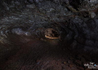 A dark Lava cave interior with uneven walls made of volcanic rock. The ground is covered in loose stones and dirt, and faint light is visible at the far end, illuminating a section of the cave. The ceiling has a rough texture, showing variations in color and sheen, indicating mineral deposits.